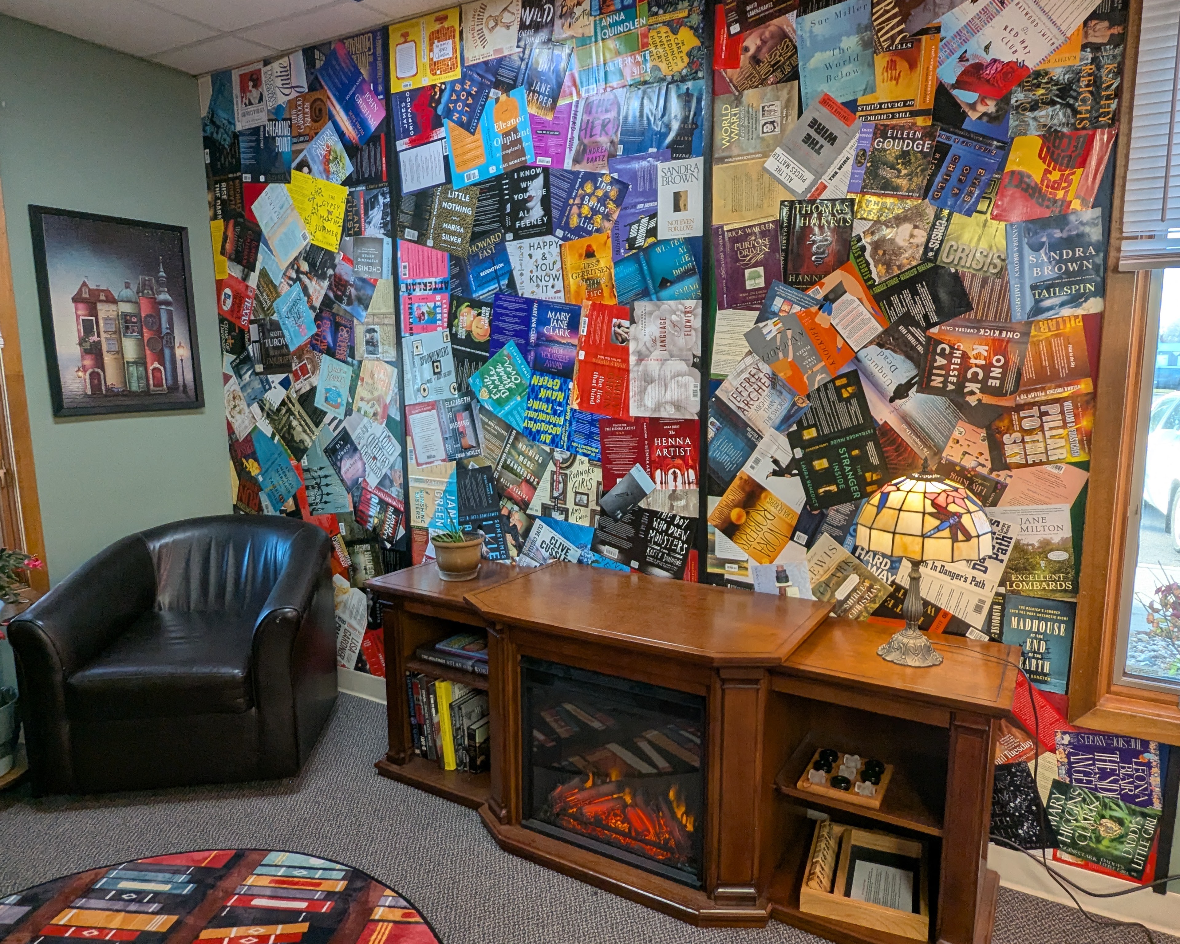 A cozy reading nook with a collage of book covers on the wall over an electric fireplace.
